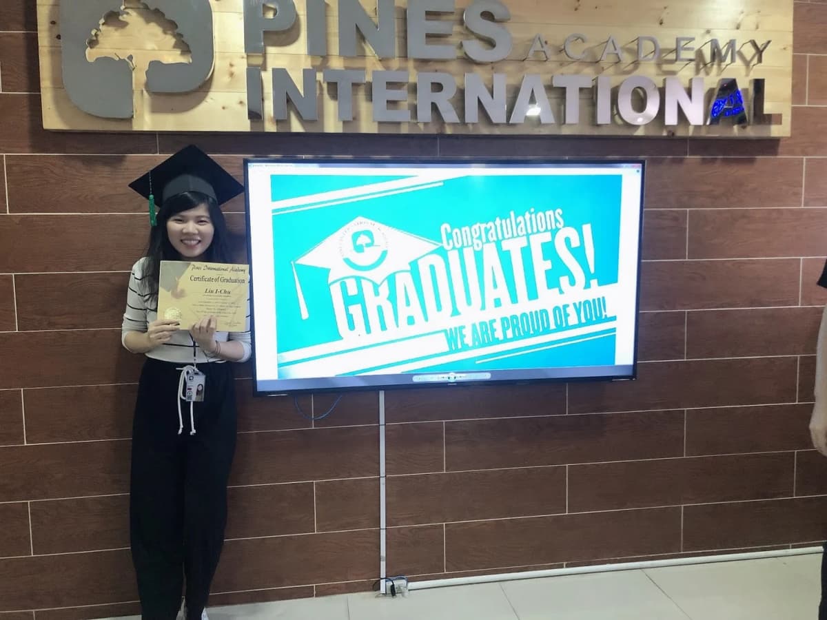Roseline in a graduation cap, holding a completion certificate in front of the PINES International Academy name wall